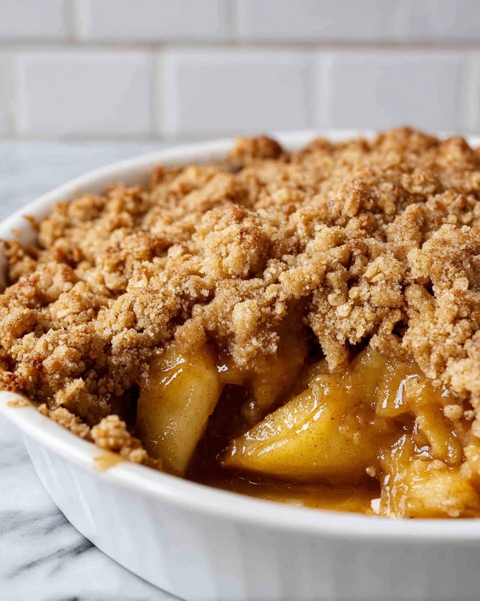A close-up image of a baked apple crumble in a round white dish, showing two main layers: a chunky, golden brown crumb topping with a rough and grainy texture on top, and underneath, visible soft, syrupy, caramel-colored apple pieces with a glossy finish. The crumble layer is thick and uneven, covering most of the filling, while some apple slices peek out near the edge. The background shows a soft white marbled surface and a white brick wall. photo taken with an iphone --ar 4:5 --v 7