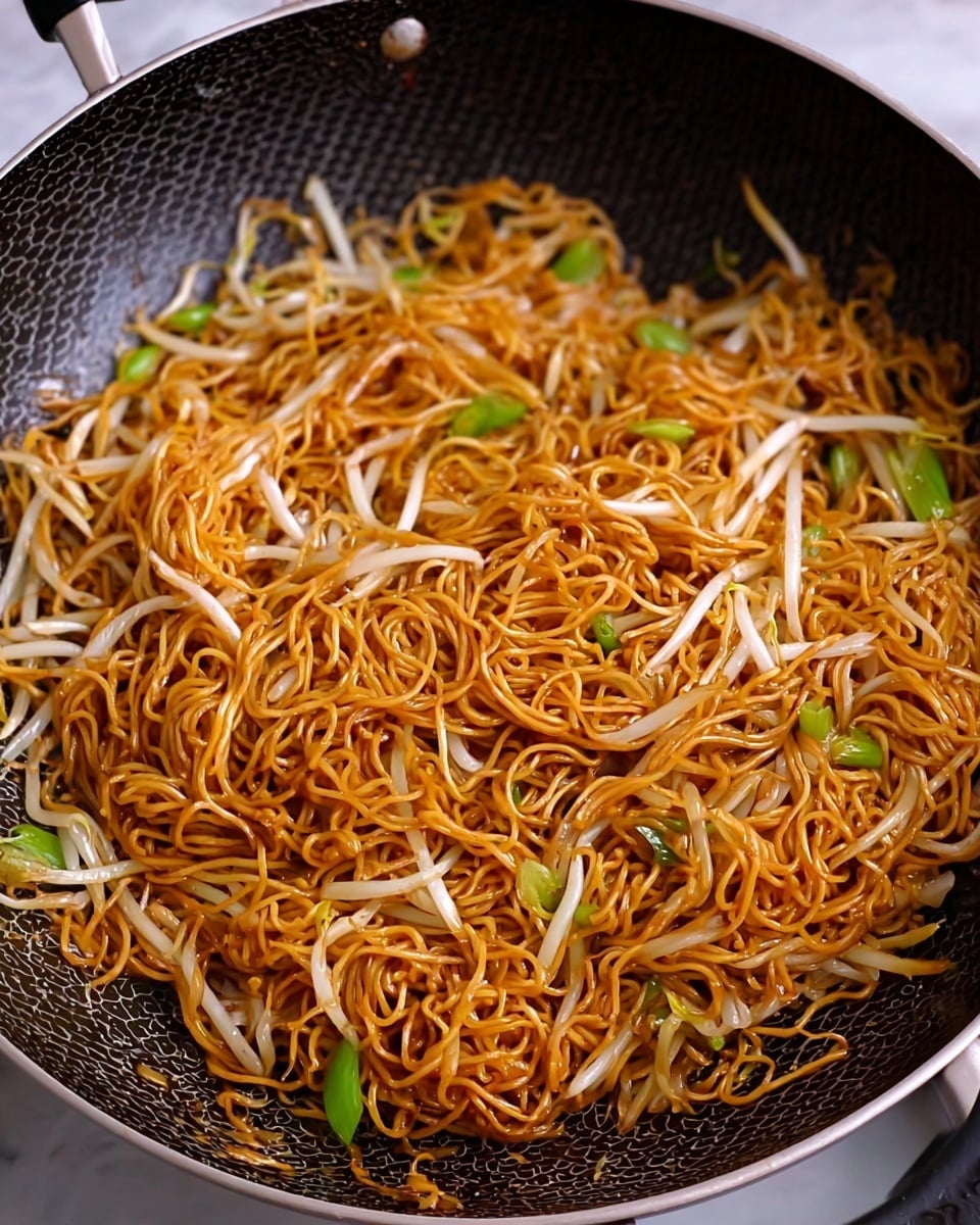 A close-up view of a pan filled with stir-fried noodles, showing one main layer of thin, curly golden-brown noodles mixed with light white bean sprouts and small green onion pieces scattered throughout. The noodles have a slightly oily texture and are evenly coated in a rich, shiny sauce. The pan's inner surface is dark gray with a hexagon pattern, providing contrast to the bright noodles. The scene is set on a white marbled texture. photo taken with an iphone --ar 4:5 --v 7