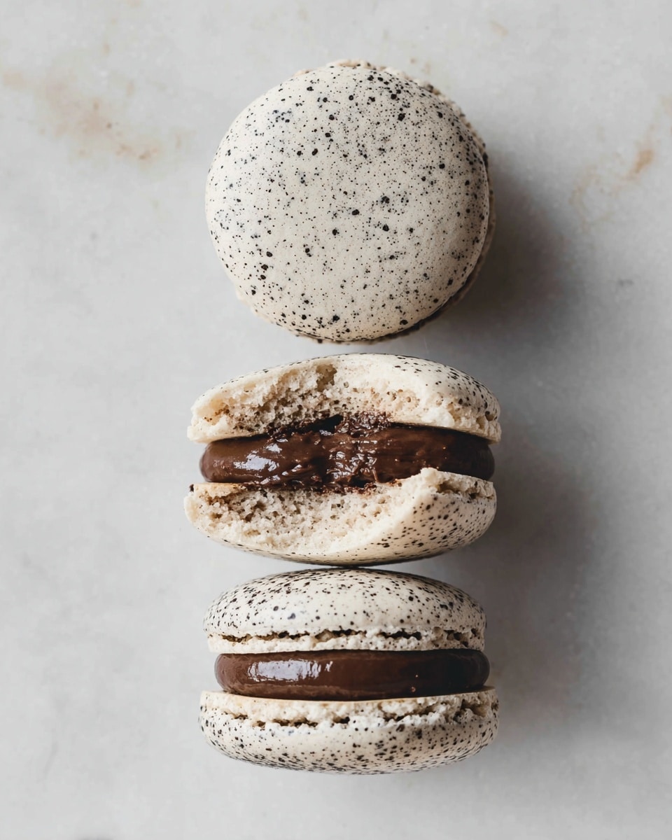 The image shows three black-speckled, off-white macarons arranged vertically on a white marbled surface. The top macaron shell is whole and smooth with tiny black specks. Below it, the matching bottom shell is open and filled with a thick, dark brown, smooth chocolate filling, clearly showing one layer of filling. At the bottom, two fully assembled macarons show two creamy shells sandwiching the rich chocolate filling inside. The texture of the shells looks slightly rough with small dots, and the filling appears glossy and dense. photo taken with an iphone --ar 4:5 --v 7