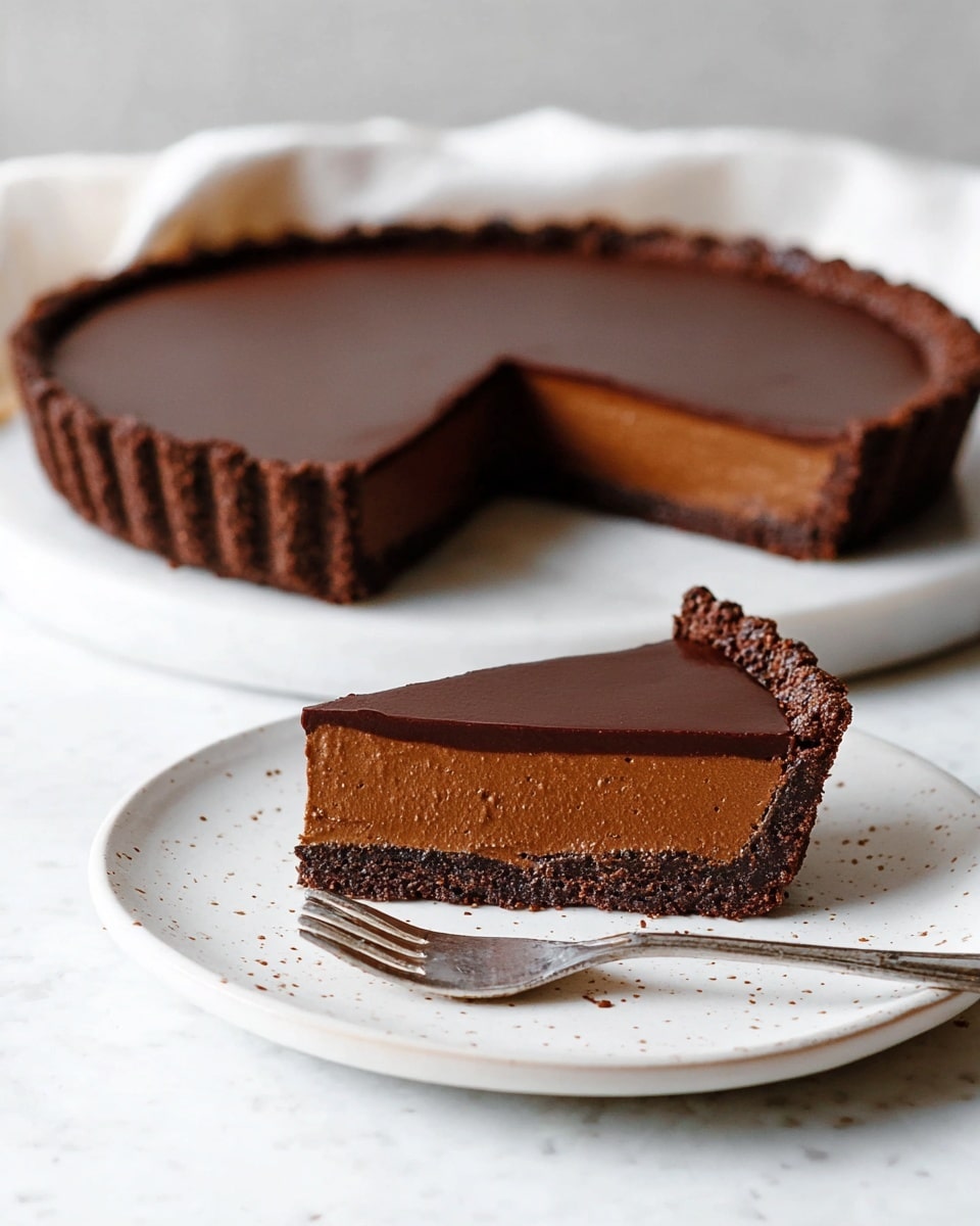 A smooth dark chocolate tart with one visible layer, consisting of a rich glossy chocolate filling that covers the entire top, set inside a thick, firm-looking chocolate crust with ridged edges. The tart is placed on a white marble board with soft grey veining, and in the blurred background, there are stacked white plates and forks along with a silver utensil resting nearby. The overall look is simple, clean, and elegant. photo taken with an iphone --ar 4:5 --v 7