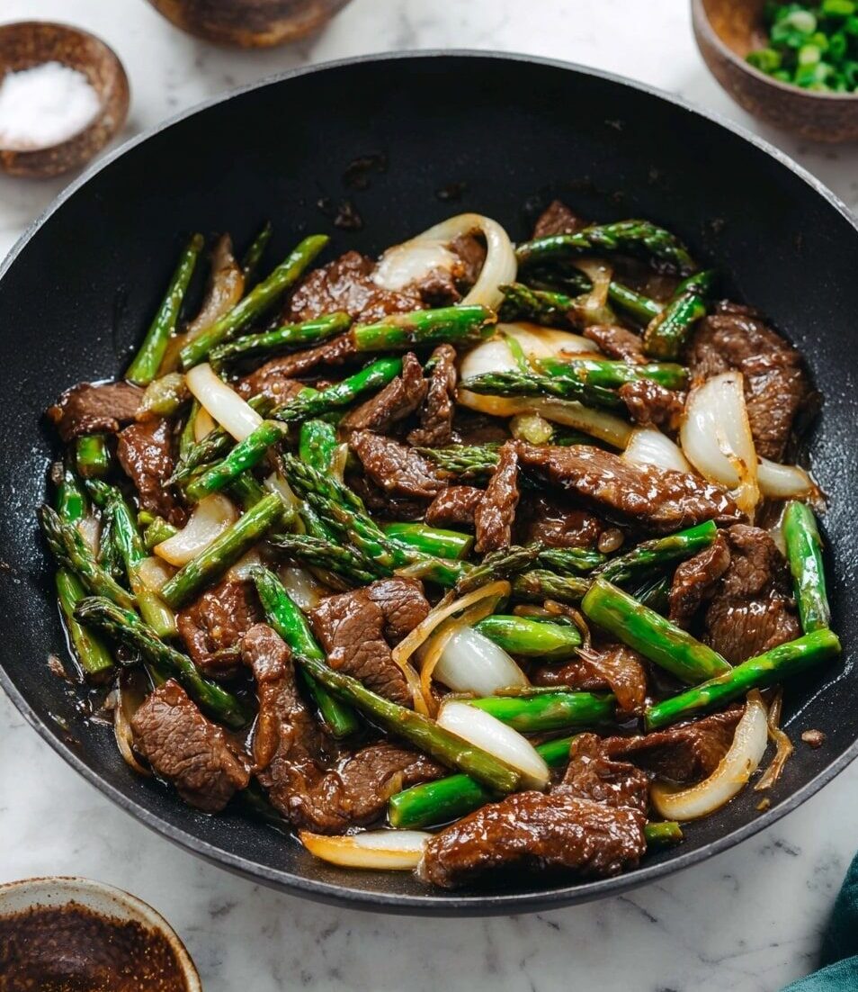 In a black wok, there is a mix of cooked beef slices with a brown glazed look, green asparagus cut into pieces, and sliced white onions. The beef has a shiny, slightly oily texture, and the asparagus looks tender but still fresh and bright green. The onions are soft and translucent, spread evenly among the other ingredients. The dish is surrounded by a few small bowls in the background, all placed on a white marbled surface. Photo taken with an iphone --ar 4:5 --v 7
