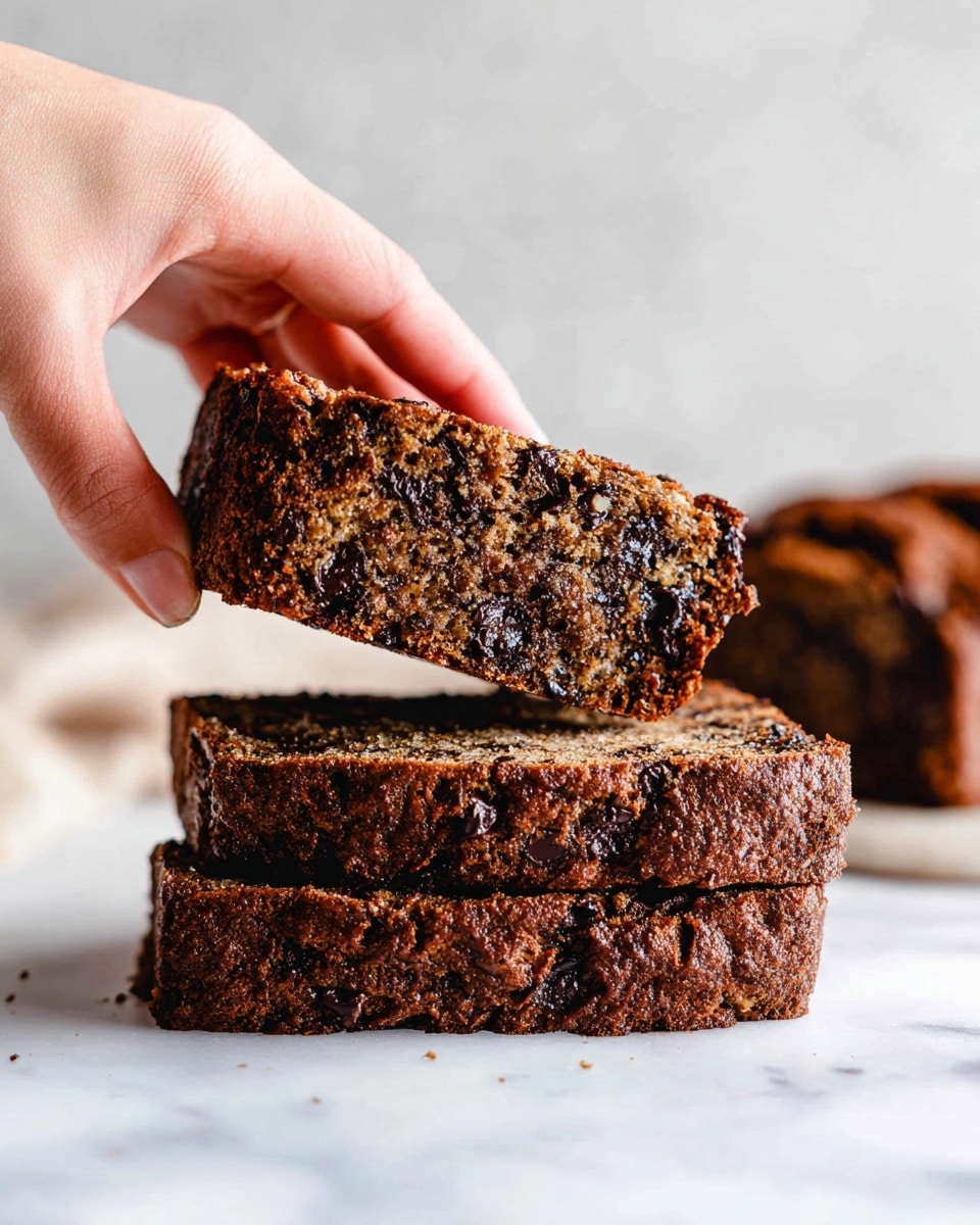 The image shows a single slice of moist banana bread lying flat on a white marbled surface. The bread has two visible layers: a darker, caramelized crust with a rough texture on the sides and top, and a lighter, soft interior speckled with small dark spots throughout. A bite is taken out of one corner, revealing the dense but crumbly texture inside. In the slightly blurred background, more slices of the same banana bread are stacked on a white cloth on a wire rack, maintaining a cozy and warm atmosphere. photo taken with an iphone --ar 4:5 --v 7