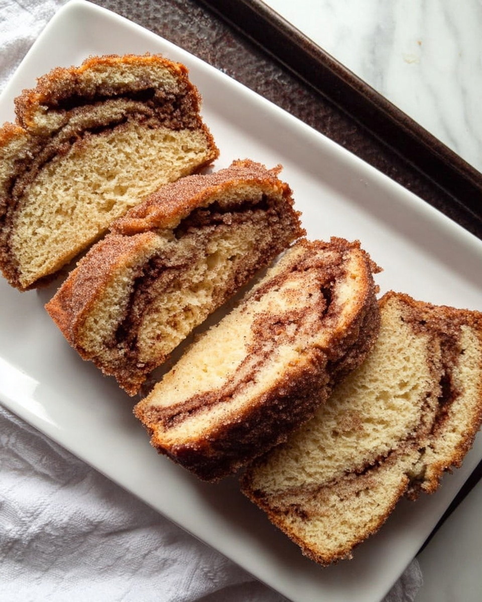 A loaf of cinnamon swirl cake on a white plate with two slices cut and laid beside it; the cake has a golden brown crust with a rough, slightly crunchy top dusted with sugar, inside is soft and light yellow with dark brown cinnamon swirls running through the middle and edges, showing a moist texture. The white plate rests on a white marbled surface. photo taken with an iphone --ar 4:5 --v 7