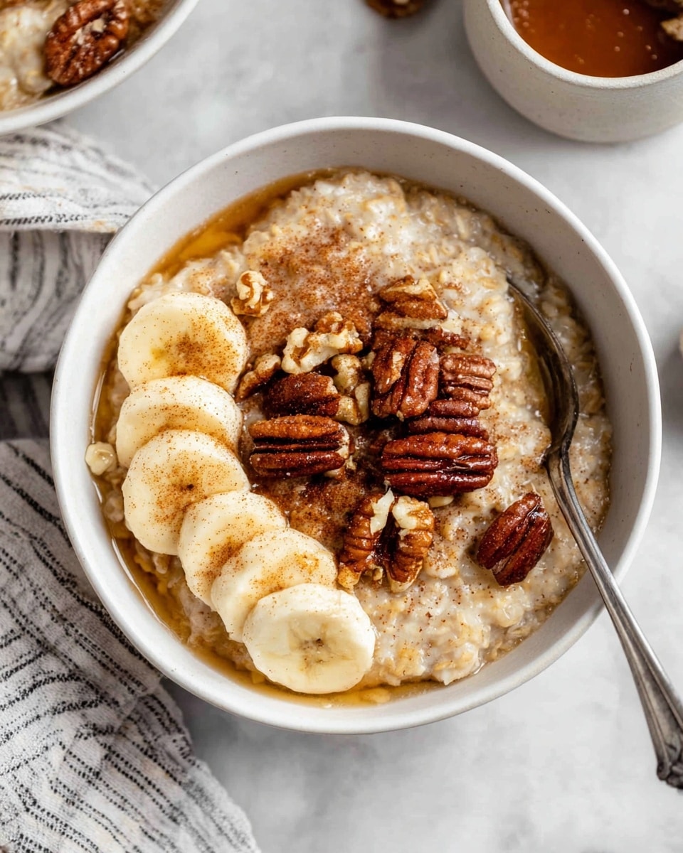 A white bowl filled with creamy oatmeal forms the base layer, topped unevenly with a golden-brown syrup that lightly pools around the edges. On one side, there are four thick, pale yellow banana slices arranged in a gentle overlap, each sprinkled softly with brown cinnamon powder. Scattered centrally over the oatmeal are large, glossy, dark brown pecan halves mixed with smaller chopped walnut pieces, adding a crunchy texture. A silver spoon rests inside the bowl on the right side, its handle extending outwards. The bowl lies on a white marbled surface with parts of other bowls and a striped cloth visible around it for a cozy setting. photo taken with an iphone --ar 4:5 --v 7
