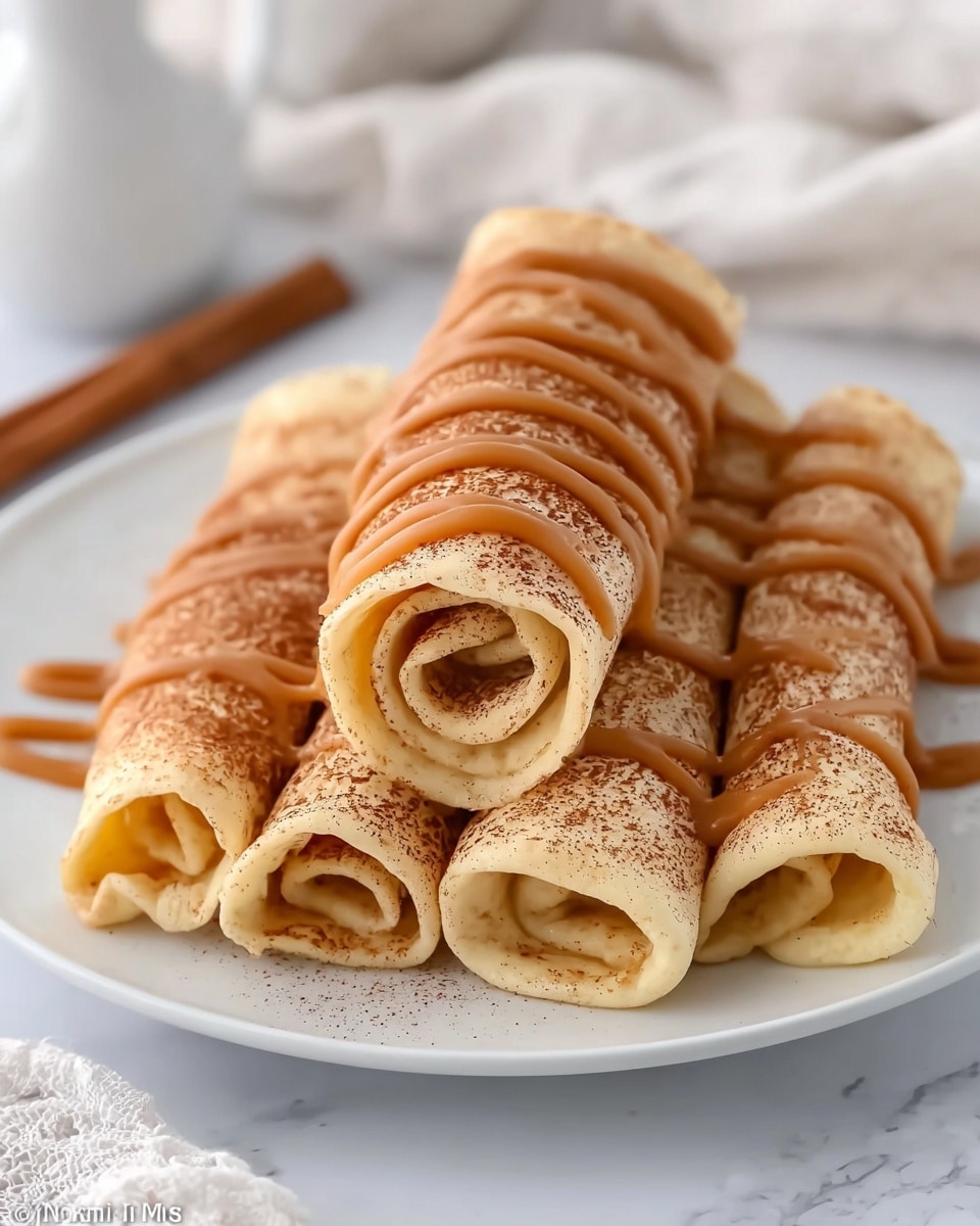 The image shows a white plate on a white marbled surface with five rolled crepes stacked in a pyramid shape. The top crepe is centered above four crepes forming the base. Each crepe is pale yellow with a light dusting of brown cinnamon powder. Caramel-colored sauce is drizzled in wavy lines over the top and sides of the crepes, creating a glossy texture and soft curves. The background is softly blurred white fabric and light-colored objects, keeping the focus on the warm, inviting crepes. photo taken with an iphone --ar 4:5 --v 7