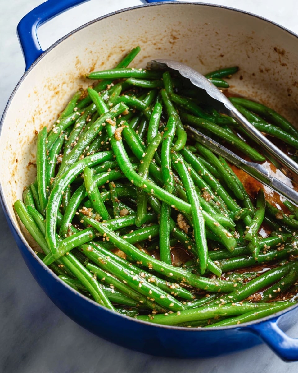 A blue pot filled with bright green cooked green beans, sprinkled with small sesame seeds and bits of garlic or seasoning, with a slight gloss over the beans showing they are well-coated in sauce, and a silver pair of tongs resting on the right side inside the pot; the pot sits on a white marbled surface. photo taken with an iphone --ar 4:5 --v 7
