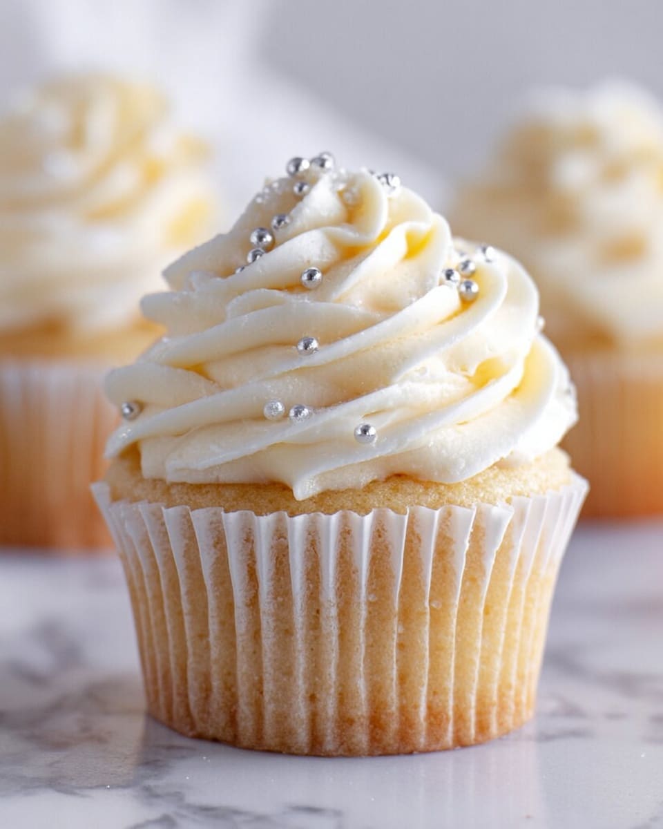 A close-up of a single cupcake with a light golden-brown base wrapped in a white paper liner, topped with a thick swirl of creamy white frosting arranged in multiple smooth, curved layers. Small shiny silver ball decorations are scattered on the frosting, adding a delicate sparkle to the soft texture. The cupcake sits on a white marbled surface with a blurred background that hints at other similar cupcakes. photo taken with an iphone --ar 4:5 --v 7
