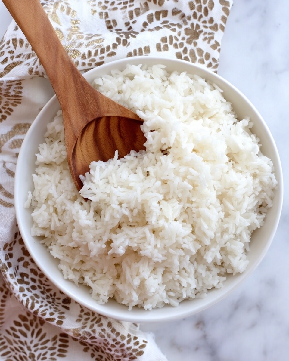 A white bowl filled with fluffy, cooked white rice showing individual grains with a soft texture, a wooden spoon resting inside the bowl, slightly buried in the rice; the bowl is placed on a white marbled surface with a white and brown patterned cloth partially visible in the background, creating a simple and clean food presentation, photo taken with an iphone --ar 4:5 --v 7