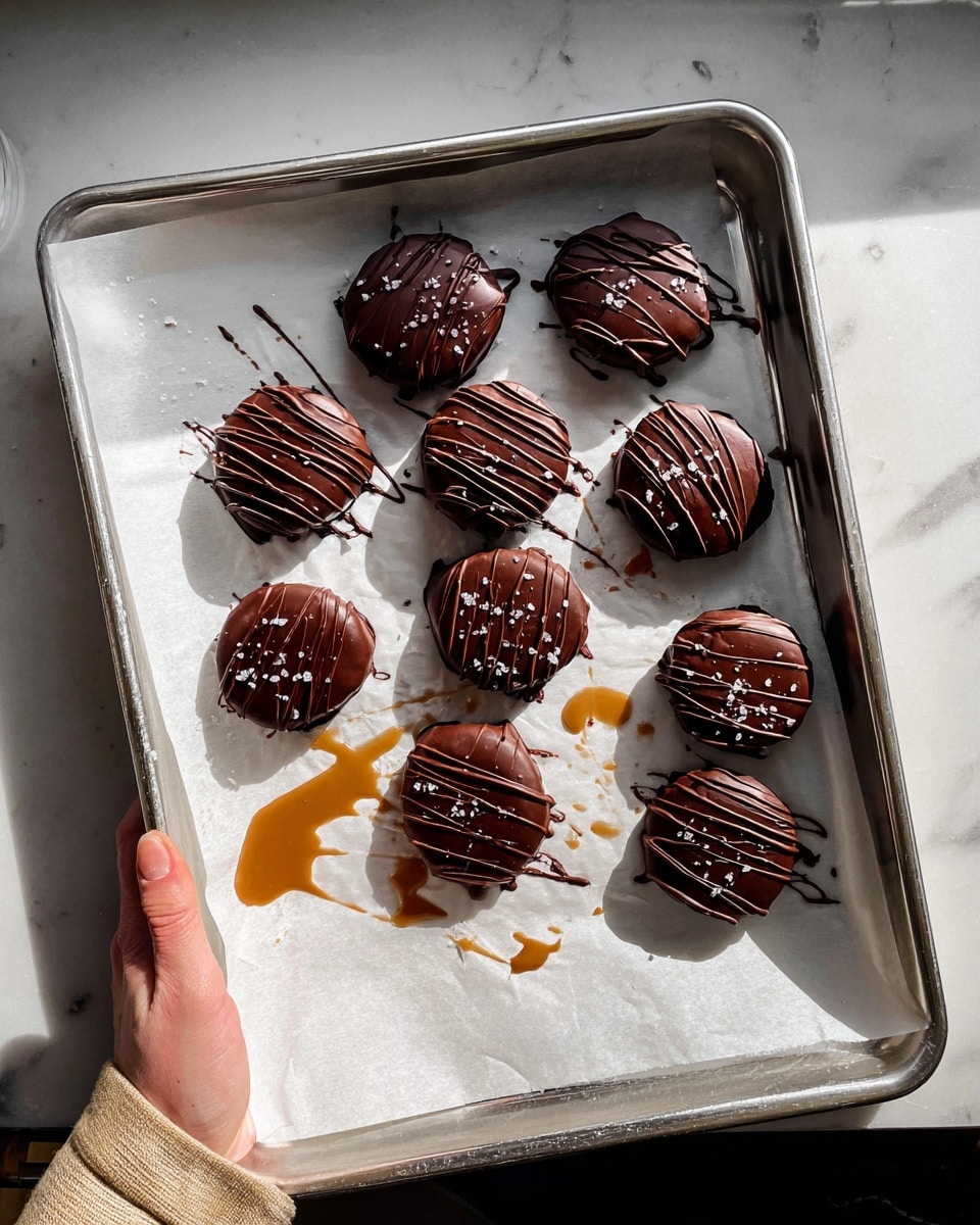 The image shows a metal baking sheet lined with white parchment paper holding nine unevenly shaped, round chocolate-covered treats. Each treat has a smooth, glossy chocolate coating with a drizzle of darker chocolate lines across the top, and a sprinkle of coarse sea salt. There are two color tones visible: some treats in a darker, almost black chocolate, and others in a lighter, milk chocolate brown. A few caramel-like drizzles are underneath and around the treats, with one larger splash near the bottom left. A woman's hand grips the right edge of the tray, held over a white marbled textured surface in bright natural light. Photo taken with an iphone --ar 4:5 --v 7