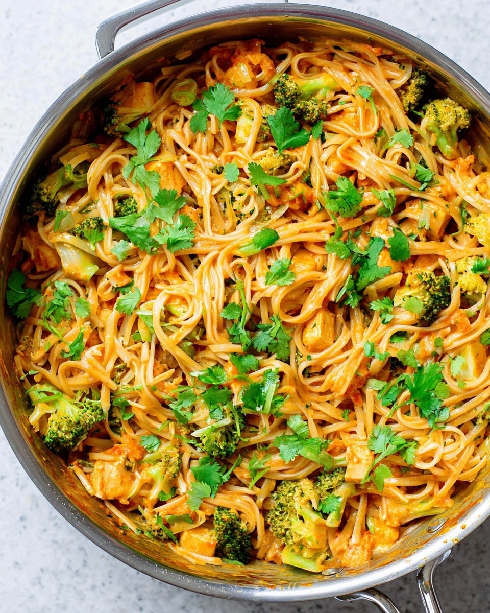 A close-up view of a pan filled with a vibrant noodle dish showing thin, light brown noodles mixed with pieces of bright green broccoli and small chunks of light orange tofu or chicken. The noodles and ingredients are coated in a creamy orange sauce, and fresh green cilantro leaves are scattered evenly on top. The pan is stainless steel and sits on a white marbled surface. The colors are bright and the texture looks smooth and creamy with some crunch from the vegetables. photo taken with an iphone --ar 4:5 --v 7