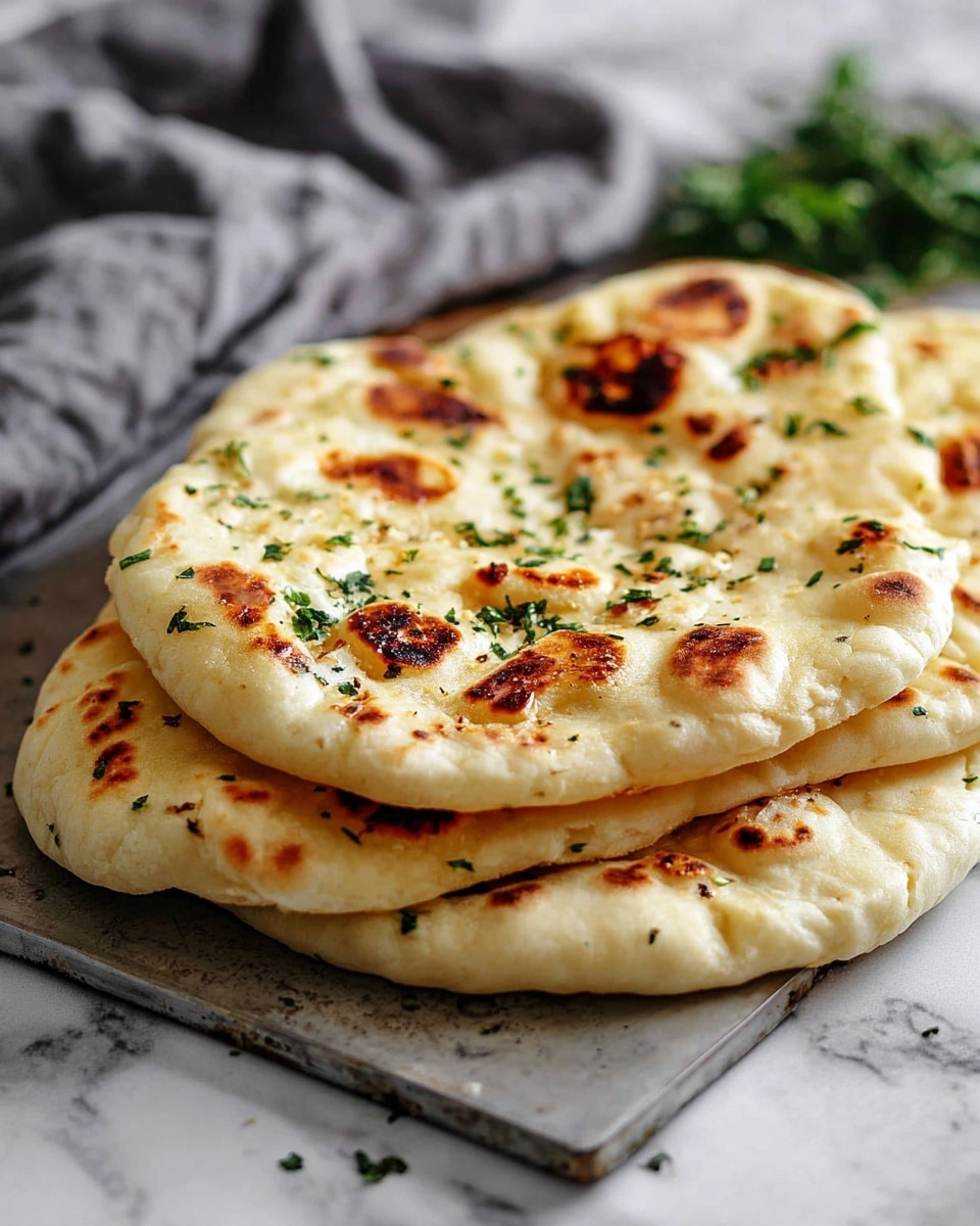 The image shows three pieces of flatbread stacked on top of each other on a metal board, placed on a white marbled surface. Each flatbread has a slightly irregular round shape with a soft and puffy texture, light golden-brown spots with some darker char marks scattered unevenly across the surface, and small green herb pieces sprinkled on top for garnish. The flatbreads appear thick and fluffy with visible air pockets and layers. In the blurry background, there is a gray cloth and some green herbs. The photo taken with an iphone --ar 4:5 --v 7