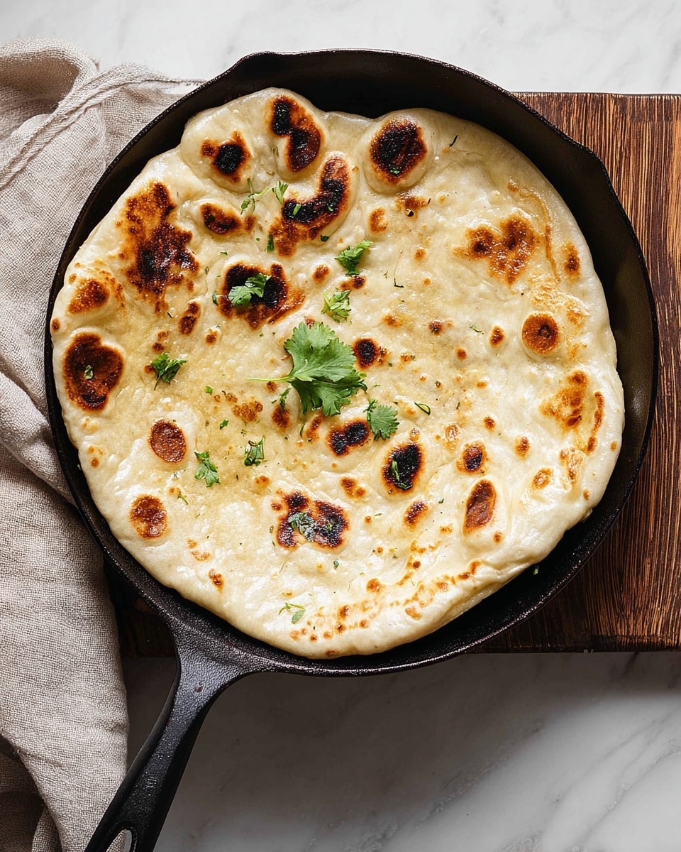 A single round flatbread with a puffy texture and browned spots is shown lying flat in a dark skillet with a long handle. The flatbread has slightly charred golden-brown patches and some lighter cream-colored areas, giving it a toasted look. There are a few small green cilantro leaves placed on top near the center, adding a fresh touch of color. The skillet is resting on a wooden board that contrasts with the white marbled background underneath. photo taken with an iphone --ar 4:5 --v 7