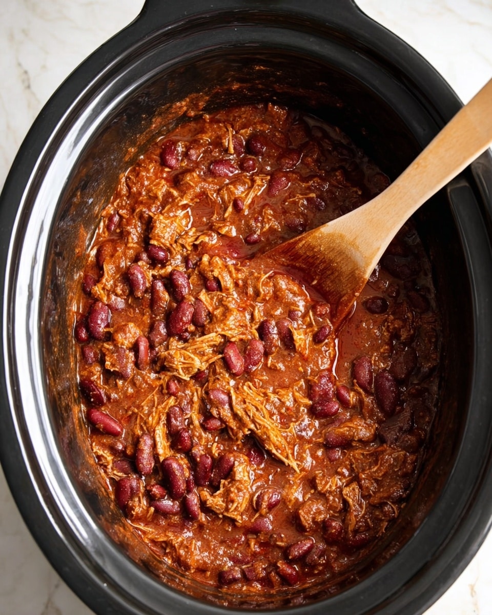 A black slow cooker filled with thick, rich chili made of shredded meat and dark red kidney beans in a deep reddish-brown sauce. A wooden spoon is partially submerged in the chili on the right side, with sauce coating the spoon and inside the cooker. The chili looks chunky and hearty with visible pieces of meat and beans mixed together. The cooker sits on a white marbled surface. photo taken with an iphone --ar 4:5 --v 7