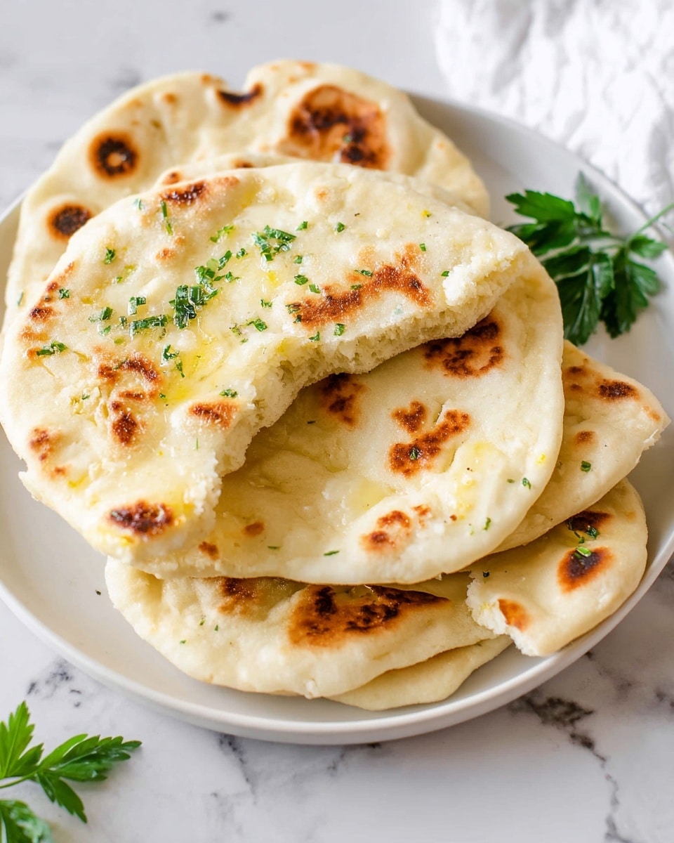 A white plate holds four pieces of naan bread stacked unevenly, showing the soft, fluffy texture of each layer. The top naan has a bite taken from it, revealing its light, airy inside with small bits of chopped green herbs and melted butter glistening on the surface. The breads are pale golden with darker brown spots where they were cooked, creating a slightly charred look. The plate is on a white marbled surface, and a few green leaves are placed around the plate to add a fresh touch. photo taken with an iphone --ar 4:5 --v 7