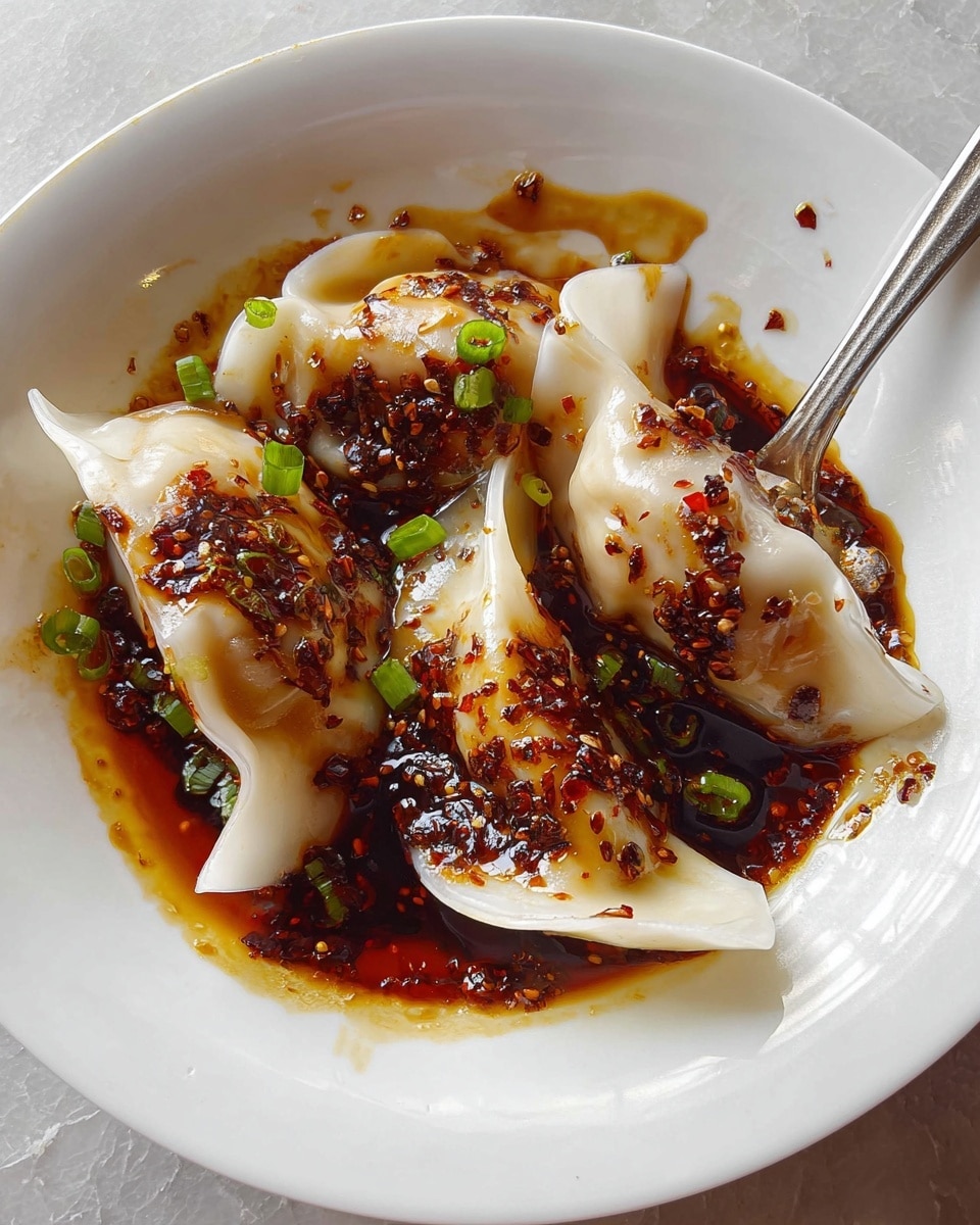 A white plate holds three dumplings with smooth, slightly translucent white skins folded into small crescent shapes, each covered with a glossy dark brown sauce that has a thick, oily texture and is speckled with tiny red chili flakes. Small green onion bits are scattered on top of the dumplings and around the sauce, adding a fresh touch of green. A silver fork rests on the right side of the plate, partially touching the dumplings and sauce. The plate sits on a white marbled texture surface. photo taken with an iphone --ar 4:5 --v 7