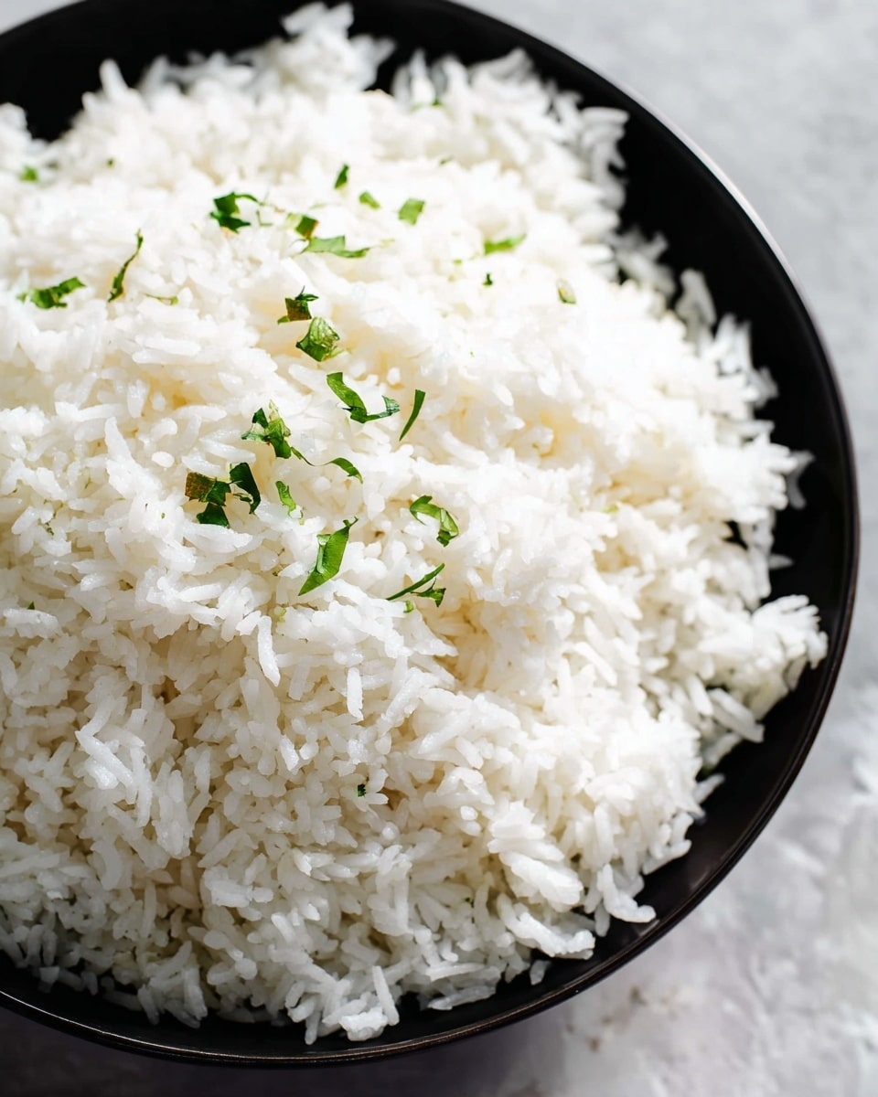 A close-up view of a black bowl filled with a single layer of fluffy white rice, scattered with a few small pieces of green herbs, placed on a white marbled texture surface. The rice grains are well separated and appear soft and light. The bowl holds the entire mound of rice, which looks freshly cooked and steaming. photo taken with an iphone --ar 4:5 --v 7