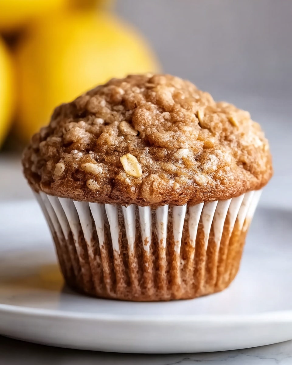 A close-up view of a single muffin with a slightly domed top textured with visible oats scattered across its rough, brown surface. The muffin’s body shows a soft, moist texture with light and darker brown spots, all wrapped in a white ridged paper liner that holds the entire baked item. The muffin sits alone on a simple white plate placed on a white marbled textured surface. In the blurred background, out-of-focus yellow fruits hint at a fresh setting. Photo taken with an iphone --ar 4:5 --v 7