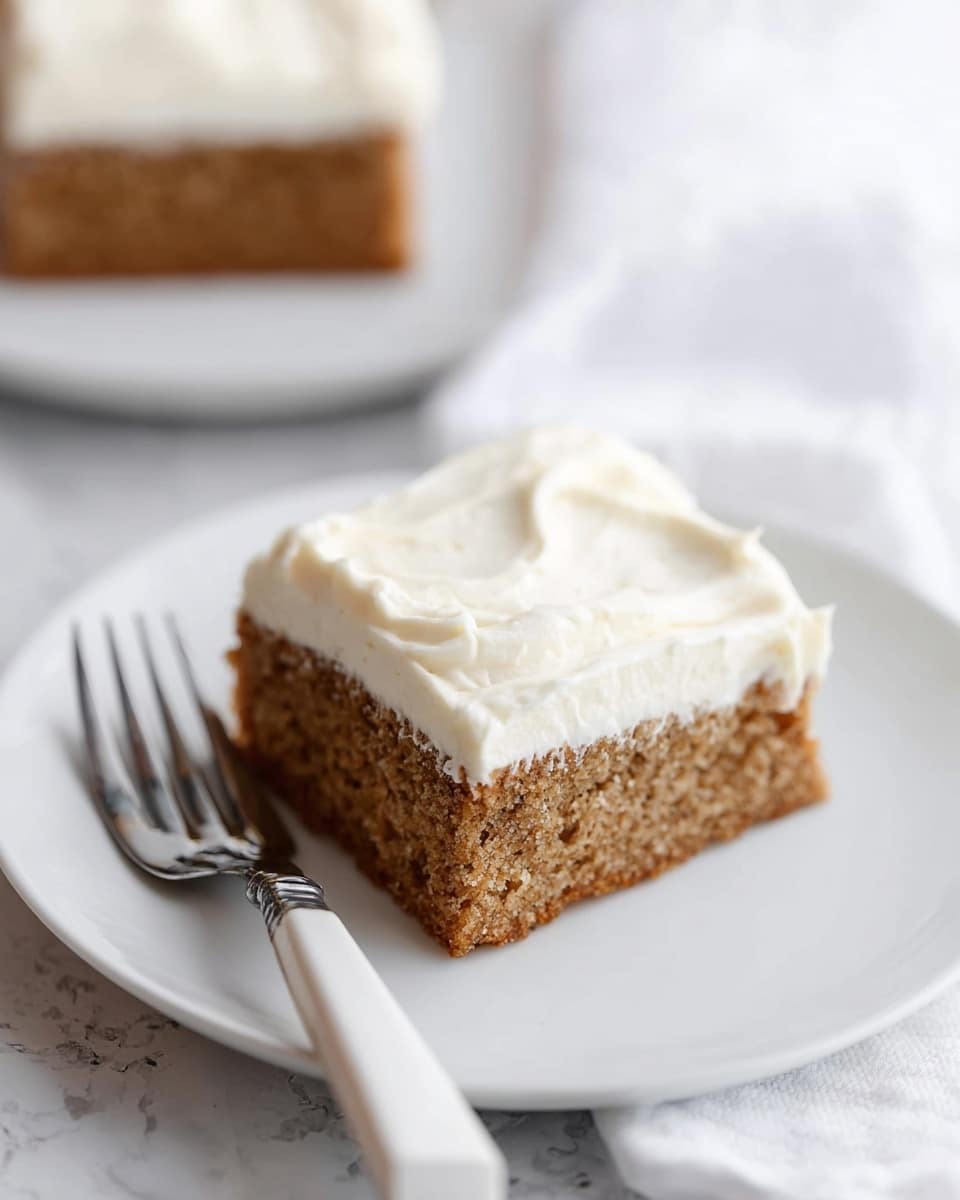 A square piece of cake with two layers is shown on a white plate placed on a white marbled surface. The bottom layer is thick, brown, and looks soft with a slightly crumbly texture. The top layer consists of a thick spread of creamy white frosting with a smooth, slightly whipped texture. Next to the cake on the plate is a white-handled fork with silver prongs. Another piece of the same cake is partially visible, blurred in the background on a similar white plate. photo taken with an iphone --ar 4:5 --v 7