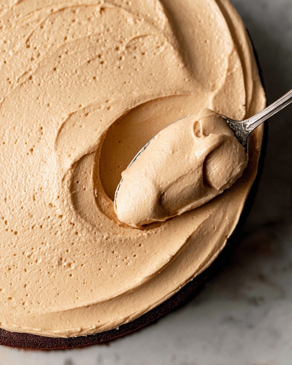 The image shows a close-up of a smooth, creamy peanut butter spread thickly inside a shallow round white bowl. The peanut butter is light brown with a slightly glossy texture, spread evenly with gentle ridges and swirls. A silver spoon scoops a rounded dollop from the center, creating a small hollow section on one side with soft peaks and a few tiny air bubbles. The bowl rests on a white marbled surface, adding a clean and bright contrast to the rich peanut butter color. photo taken with an iphone --ar 4:5 --v 7
