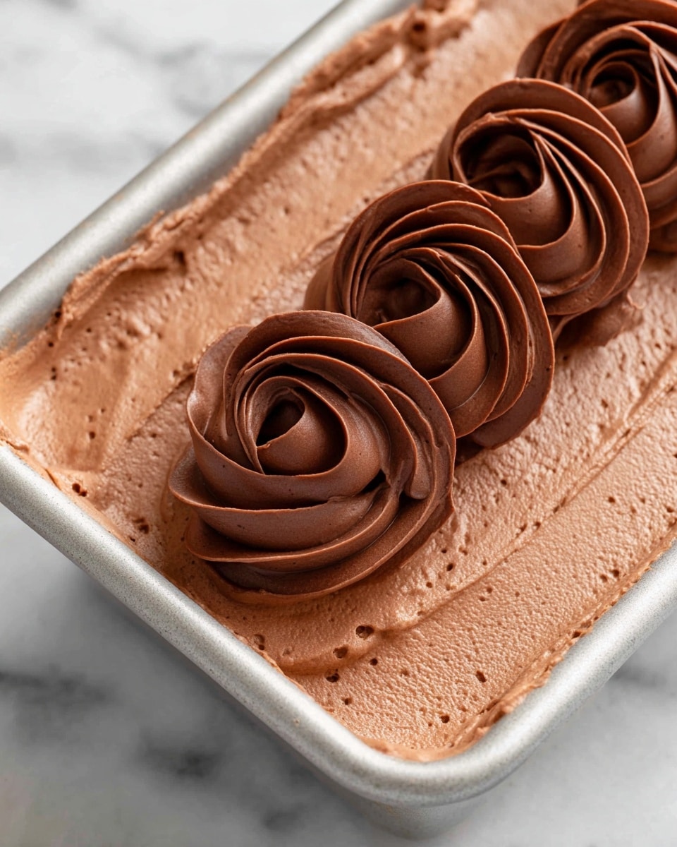The image shows a close-up view of a creamy chocolate dessert in a rectangular metal baking pan with rounded corners. The dessert is smooth and has one thick base layer of light brown chocolate mousse with a silky texture, topped with a neat row of darker brown chocolate piped in a swirl pattern, forming a series of connected loops placed right in the center, stretching lengthwise. The background is a white marbled surface. Photo taken with an iphone --ar 4:5 --v 7