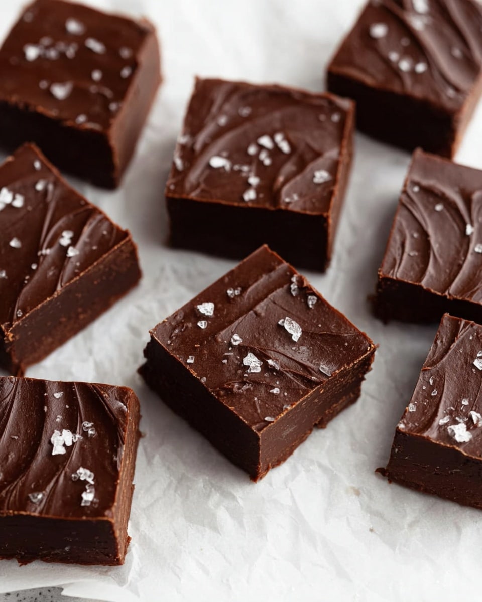 The image shows several square pieces of dark chocolate fudge arranged closely on white parchment paper over a white marbled surface. Each piece has a smooth, shiny top layer with slight swirled texture and is sprinkled with small flakes of sea salt. The fudge layers are thick and rich, appearing dense and creamy with a uniform dark brown color throughout. No other objects or utensils are seen, focusing only on the fudge squares. photo taken with an iphone --ar 4:5 --v 7