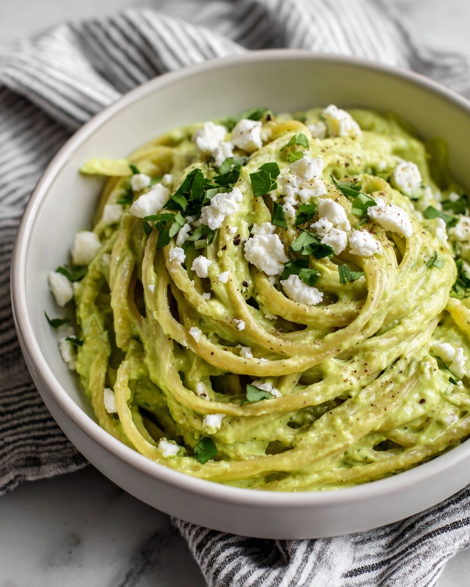 A close-up of creamy green pasta in a white bowl, showing a generous layer of smooth, light green sauce coating long noodles that twist and overlap. Small white crumbles of cheese are scattered on top, with bright green herb pieces adding a fresh touch. The bowl sits on a white marbled surface, and a striped cloth can be seen blurred in the background. photo taken with an iphone --ar 4:5 --v 7