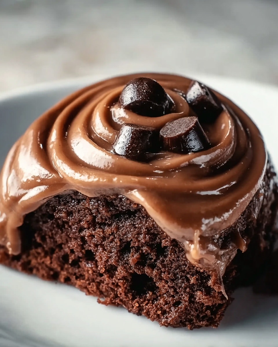 The image shows a close-up of a rich chocolate cake slice topped with a thick swirl of smooth, creamy milk chocolate frosting. On top of the frosting, there are three shiny dark chocolate chunks placed unevenly, adding texture and depth. The cake itself is dark brown, moist and dense, contrasting with the lighter brown frosting. The dessert sits on a white plate, placed on a white marbled surface. The focus is sharp on the cake and frosting, highlighting the glossy shine and the soft texture. photo taken with an iphone --ar 4:5 --v 7