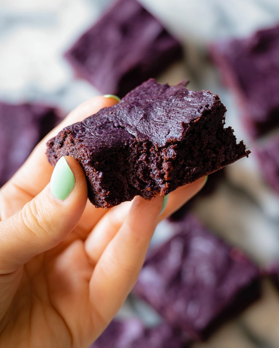 A close-up of a woman's hand holding a square-shaped piece of deep purple brownie with a slightly cracked, shiny top layer and a soft, moist, and dense inner texture visible from a bite taken out of one side. The background shows several more purple brownie pieces blurred out on a white marbled surface. The woman's thumb has a smooth, light green nail polish. photo taken with an iphone --ar 4:5 --v 7