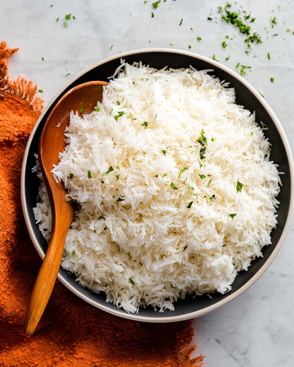 A large round white bowl is full of fluffy cooked white rice, with individual grains visible and slightly separated. The rice is sprinkled lightly with small green herb pieces on top and mixed in, adding a touch of color. A wooden spoon with a curved handle rests inside the bowl, partially buried in the rice on the left side. The bowl sits on a white marbled textured surface with a textured orange cloth in the upper left corner and scattered small green herbs nearby, enhancing the fresh look. Photo taken with an iphone --ar 4:5 --v 7