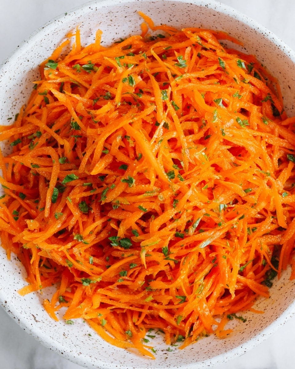 This image shows a large bowl full of finely shredded bright orange carrots mixed with small bits of green herbs scattered evenly throughout. The carrots have a fresh and slightly wet texture, suggesting they are dressed with a light sauce or vinaigrette. The bowl is white with small dark specks, and the shredded carrots fill it almost to the top, creating a vibrant, colorful mix. The background is a white marbled texture. photo taken with an iphone --ar 4:5 --v 7