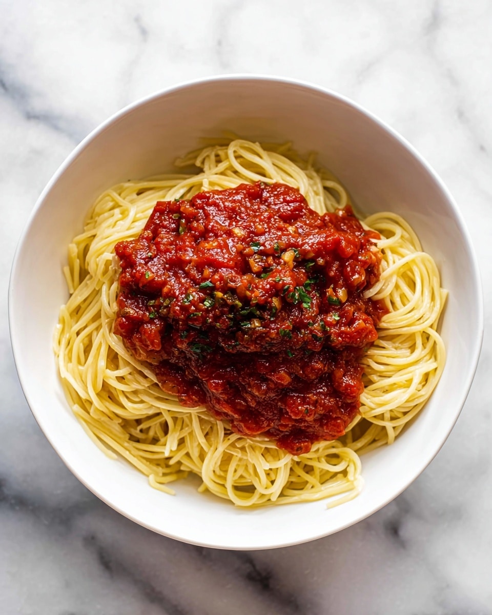 A white bowl holds a simple dish of spaghetti with tomato sauce. The bottom layer is a neat pile of light yellow cooked spaghetti pasta, forming a smooth nest. On top, a thick layer of rich red tomato sauce with visible chunks and small green flecks is spread generously, covering the center of the pasta. The bowl sits on a white marbled surface. photo taken with an iphone --ar 4:5 --v 7