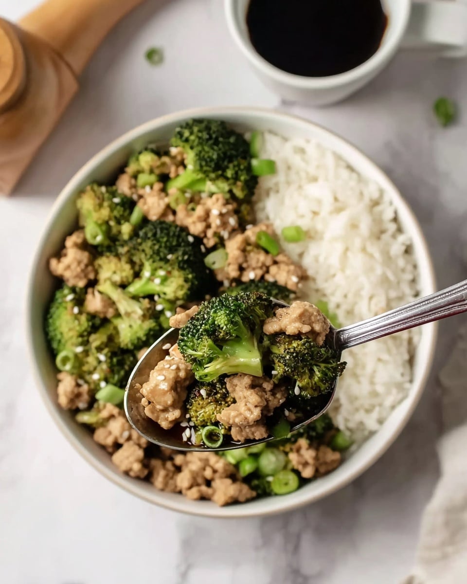 A white bowl sits on a white marbled surface filled halfway on the right with light fluffy white rice and the left half with cooked bright green broccoli pieces mixed with small light brown ground meat chunks, topped with small green onion pieces and white sesame seeds. In the foreground, a silver spoon holds a close-up of the broccoli and meat mix, highlighting the vibrant green and soft brown texture. In the background, a white cup filled with dark soy sauce and a wooden pepper grinder are slightly out of focus. Photo taken with an iphone --ar 4:5 --v 7