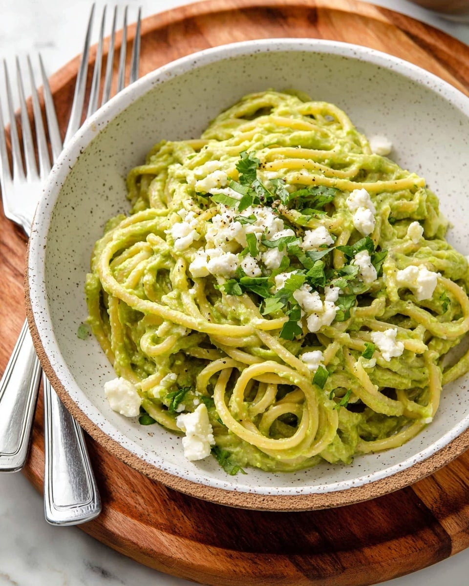A white speckled bowl filled with thick spaghetti pasta coated evenly in a creamy green sauce, likely avocado or pesto, creating a smooth, rich texture. Scattered on top are small clumps of soft white cheese and sprinkled fresh green herbs, adding contrast in both color and texture. The bowl sits on a round wooden board with two silver forks resting beside it, all set against a white marbled textured surface. photo taken with an iphone --ar 4:5 --v 7