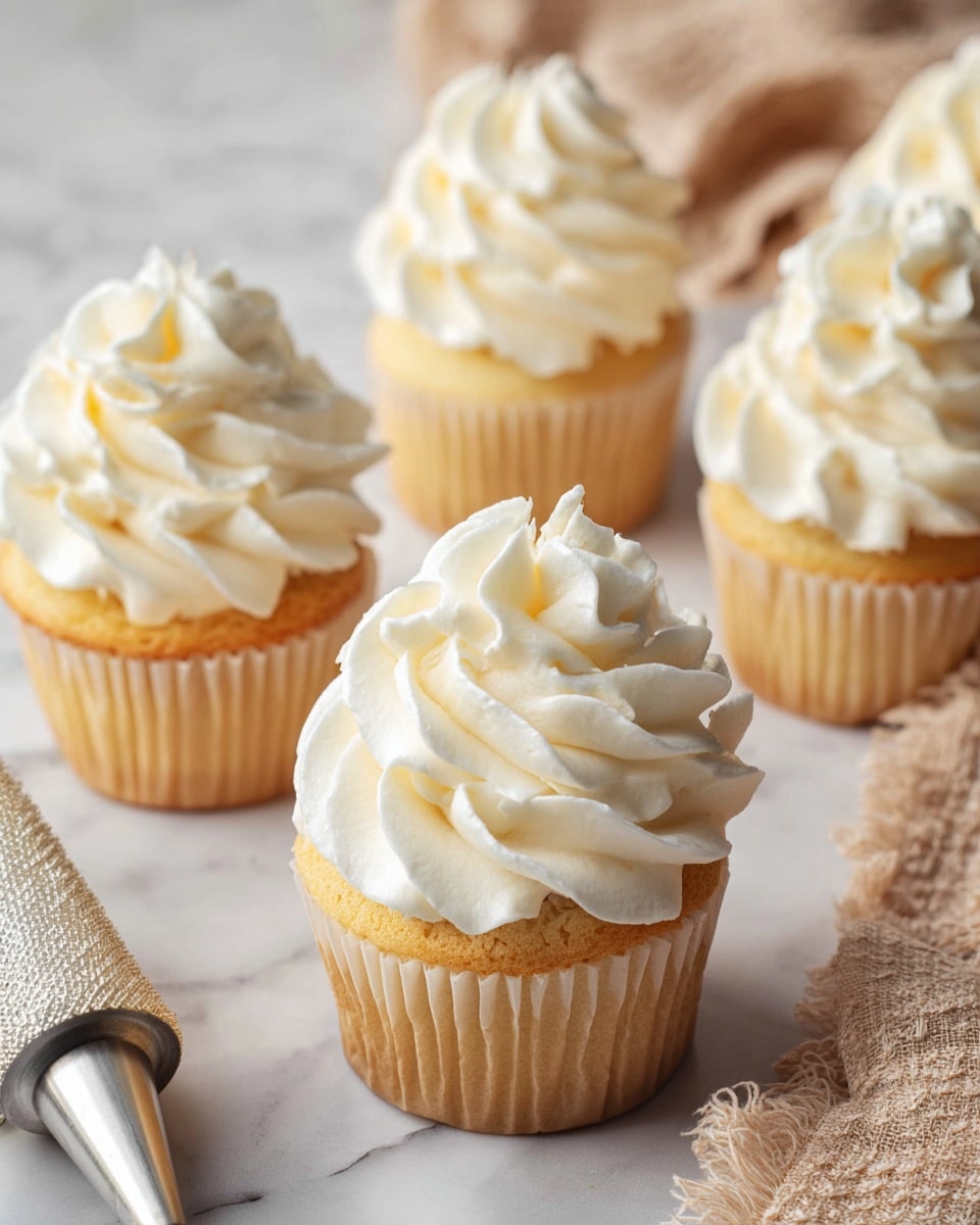 This image shows five vanilla cupcakes with tall swirls of white buttercream frosting piped on top. Each cupcake has one thick layer of golden yellow cake visible beneath the creamy, fluffy frosting. The frosting is smooth and soft with slight peaks and curves, decorating the top with large, flowing swirls. The cupcakes sit on a white marbled surface with a silver piping tip and a beige textured cloth nearby, adding a cozy baking vibe. The focus is on the cupcake in the center, showing the frosting’s texture clearly. photo taken with an iphone --ar 4:5 --v 7