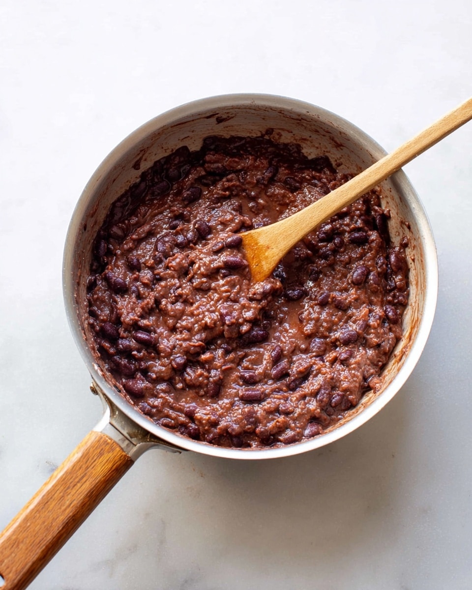 A silver pan with a wooden handle contains a thick mixture of dark reddish-brown chunky beans. A wooden spoon with a light brown handle is partly stirring the mixture, showing the soft and slightly mashed texture of the beans. The pan sits on a white marbled surface, with no other items around. The lighting is bright and natural, highlighting the rich color and moist texture of the bean dish. photo taken with an iphone --ar 4:5 --v 7