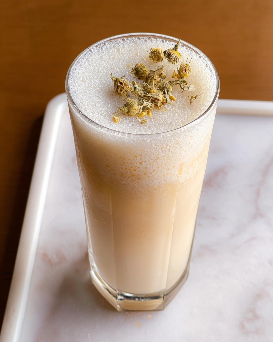 A tall, clear glass filled with a creamy light beige drink topped with a frothy foam layer. On top of the foam, there is a small cluster of dried light brown and pale yellow flowers resting gently. The glass is placed on a white marbled surface. photo taken with an iphone --ar 4:5 --v 7