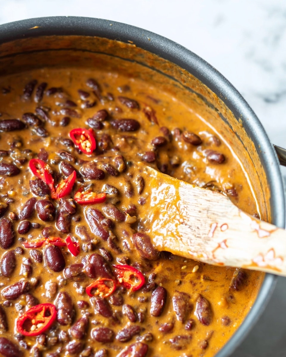 The image shows a thick stew with three visible layers in a pot. The base layer is a creamy orange sauce, covering almost the entire pot. On top of that, there are dark brown kidney beans, evenly mixed throughout the stew. Scattered on the surface are thin red chili slices, adding a bright red color contrast. A wooden spatula with sauce stains is inserted into the stew, lifting a part of it, showing the texture of the beans and sauce sticking together. The background is a white marbled texture. photo taken with an iphone --ar 4:5 --v 7
