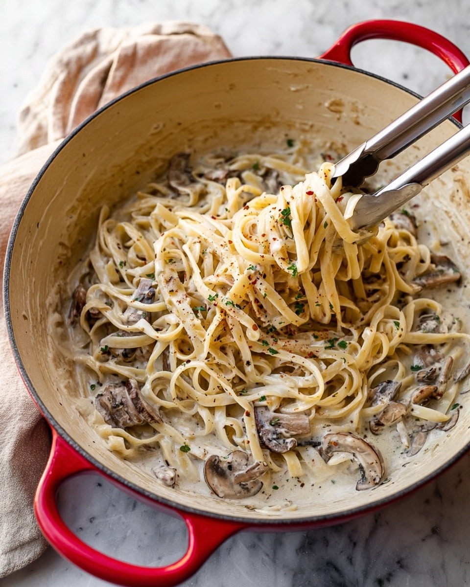 A close-up of a white pot with red handles filled with creamy mushroom pasta. The dish has one main layer of long, flat pasta strands mixed with sliced brown mushrooms in a thick white sauce speckled with herbs and spices. Silver tongs are lifting some pasta from the bottom right side of the pot. The pot sits on a white marbled surface with a beige cloth in the background. photo taken with an iphone --ar 4:5 --v 7