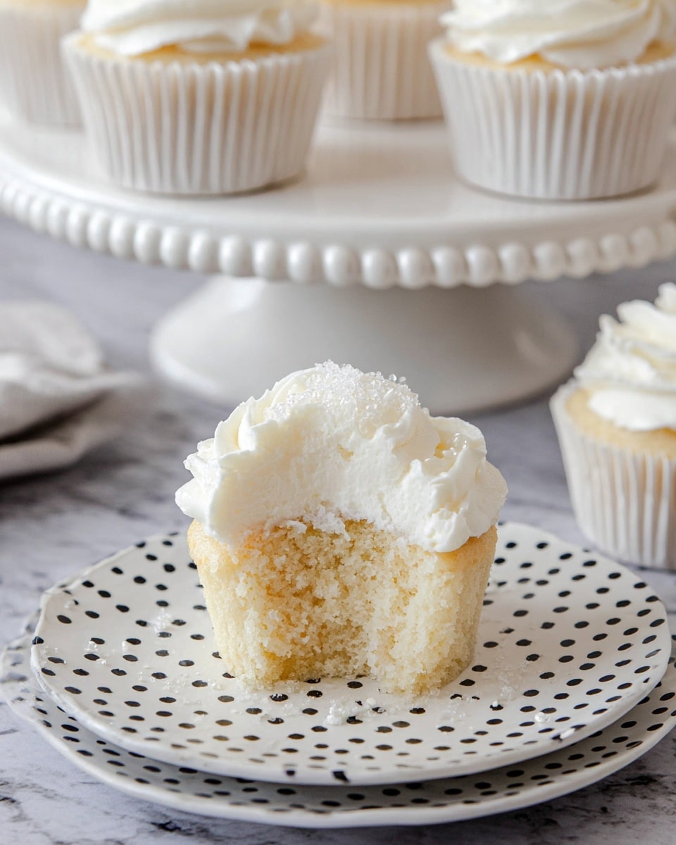 A close-up of a white cupcake with one bite taken out, showing its soft, light beige crumb texture inside; it has a thick layer of fluffy, white frosting on top, swirled with some small sugar crystals sprinkled over it. The cupcake sits on a white plate with black polka dots, which rests on a white marbled textured surface. In the background, there are more cupcakes in white paper liners arranged on a white cake stand with a round beaded edge, slightly out of focus for depth. Photo taken with an iphone --ar 4:5 --v 7
