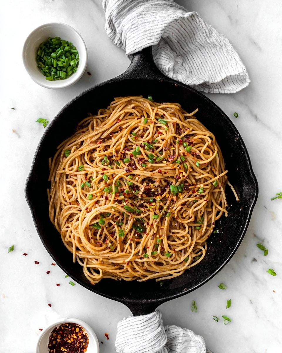A black cast iron skillet filled with a single layer of cooked spaghetti noodles coated in a light brown sauce, scattered with small bright red chili flakes and chopped fresh green chives on top, sitting on a white marbled surface. The skillet handle is wrapped with a white cloth with thin black stripes. Above the skillet, there are two small white bowls, one filled with chopped green chives and the other with red chili flakes. Some chive pieces are scattered around the skillet on the white marbled surface. photo taken with an iphone --ar 4:5 --v 7